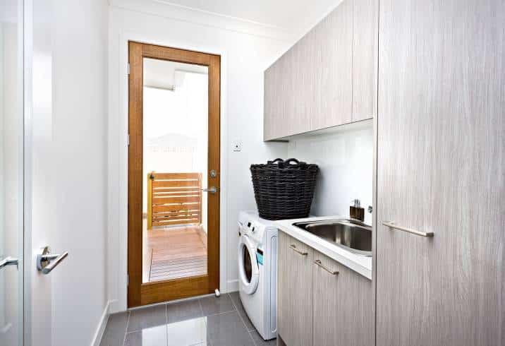 Modern laundry room near a wooden door with a glass panel closed. Grey cupboards on the wall, black basket made in rattan on the washing machine