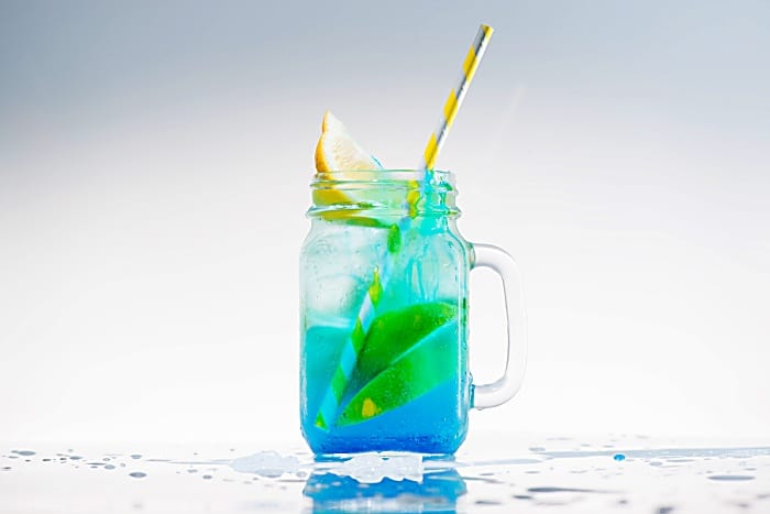 mason jar of cold blue fresh lemonade with lime, lemon and yellow drinking straw on light background