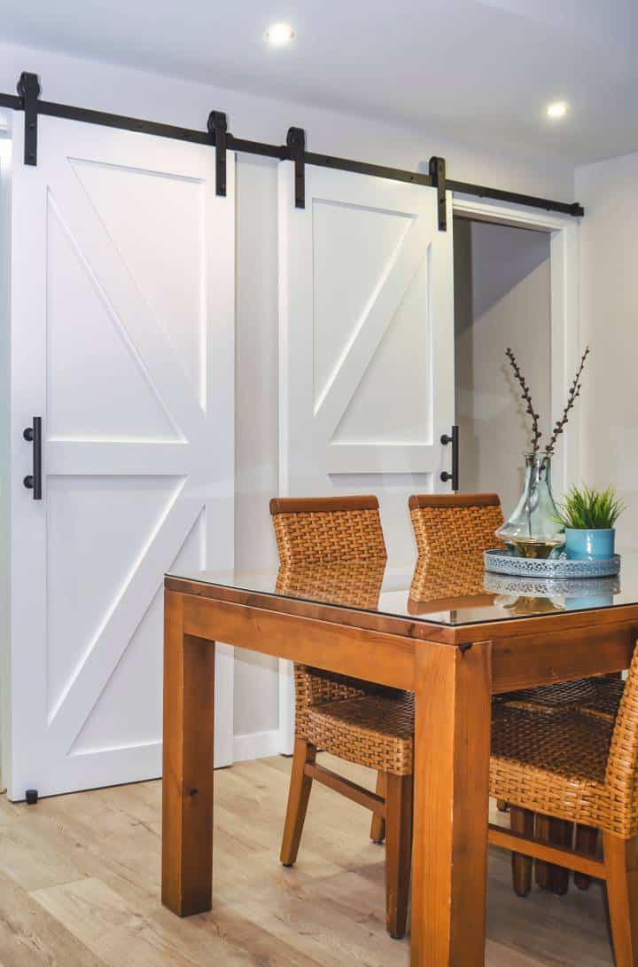 Bright dining room with a wooden table and rattan chairs. White sliding barn doors in the background.