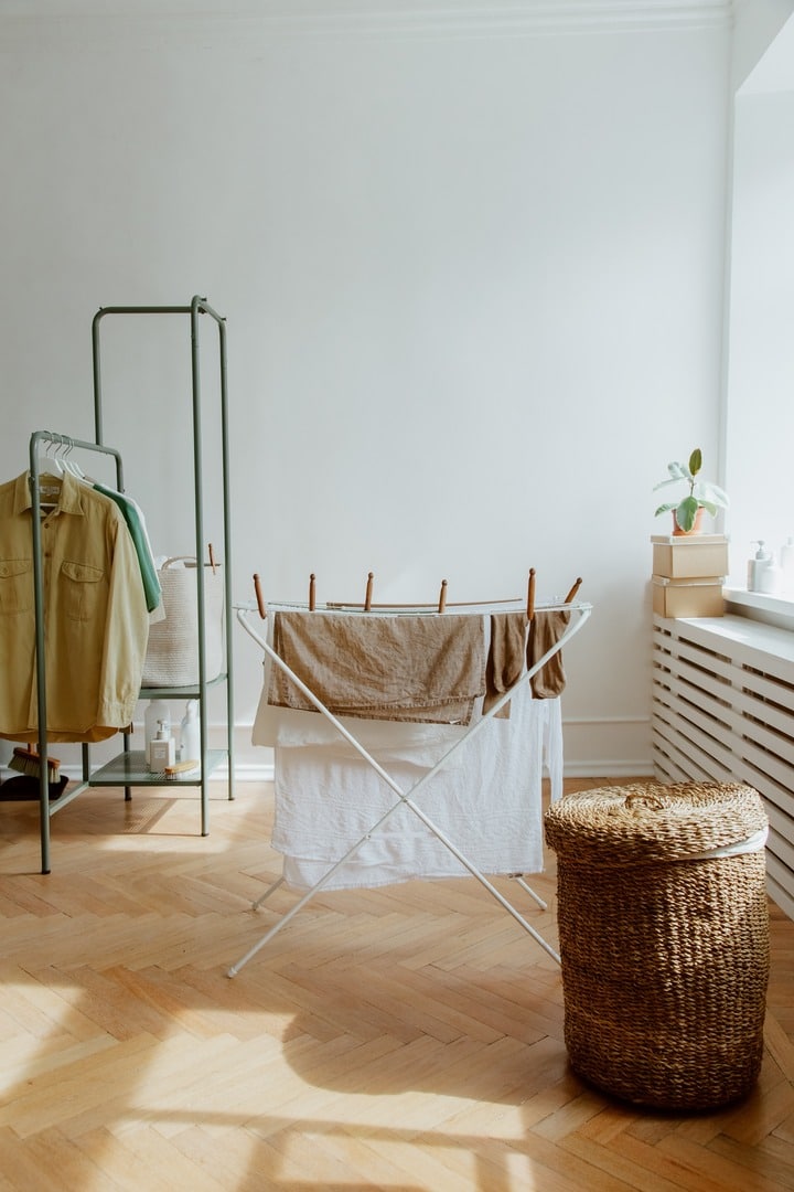 Lidded wicker laundry hamper near a laundry rack with hanging towels and socks. Sunlight shines on the wooden floor, with shadows from the windows