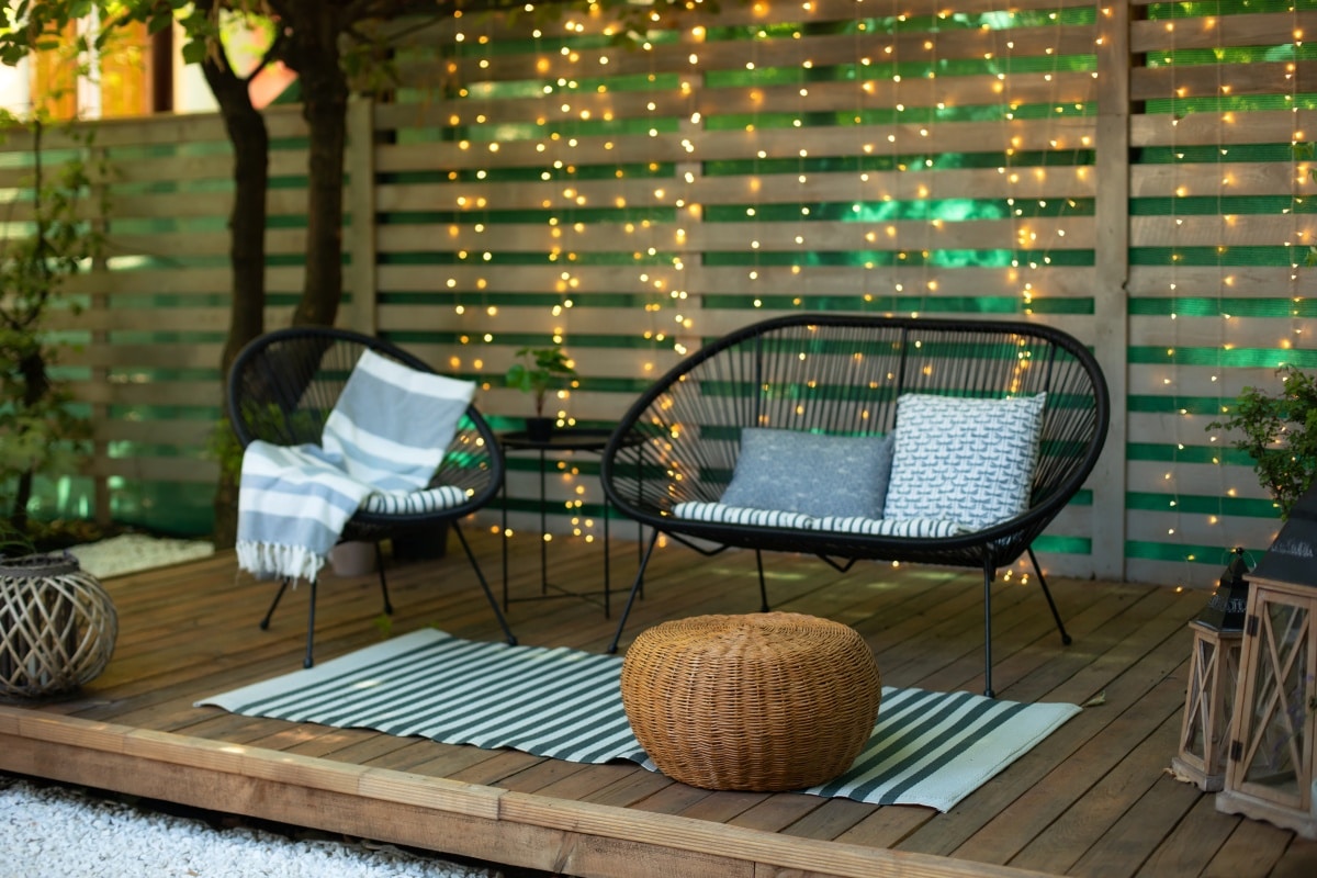 Terrace with plants, wooden wall and table, comfortable sofa, armchair and lanterns