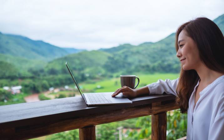 Smiling female accountant typing on laptop while sitting on balcony with mountains and green nature, enjoying remote work 