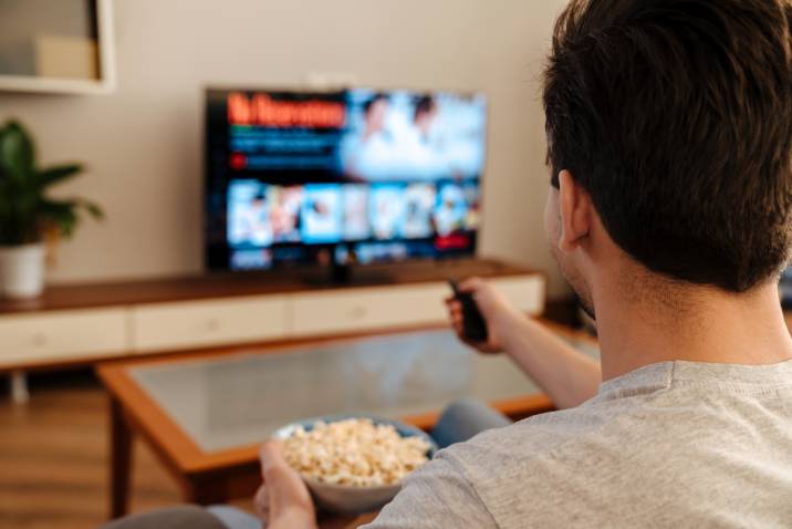 man watching TV and eating popcorn on a couch, house sitting side hustle for lazy people