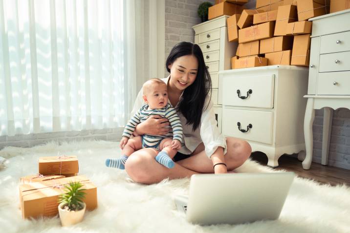 Beautiful young mother with baby boy, using laptop and preparing boxes of crafts to sell