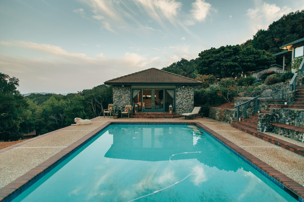 stone poolhouse with shingled roof in front of a pool