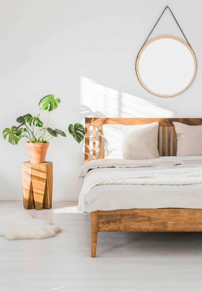 Bedroom with wooden furnishings. Monstera plant on a tree trunk nightstand and a round mirror above the bed, on a white wall in a sunlit bedroom interior