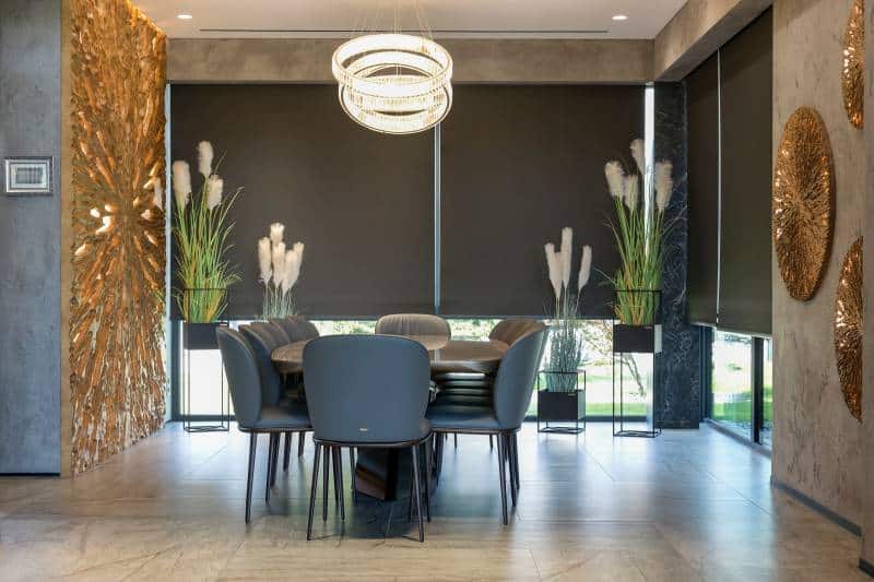 Automatic roller blackout shades on large windows in dining room. Modern interior with green plants and wood decor panels on the wall 
