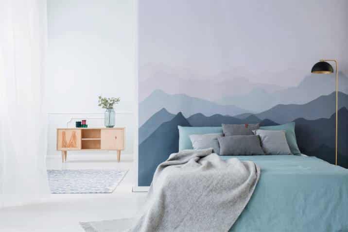 Blue bed with grey blanket against mountain wall mural. Simple bedroom interior with wooden cupboard