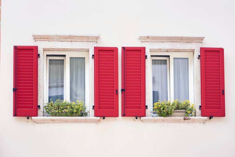 Two Italian windows on the white wall facade with open red colour classic shutters and flowers on the windowsill