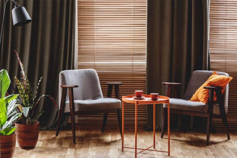 Curtains over blinds in a living room interior. Vintage armchairs, orange coffee table with two cups and plants standing by the window 
