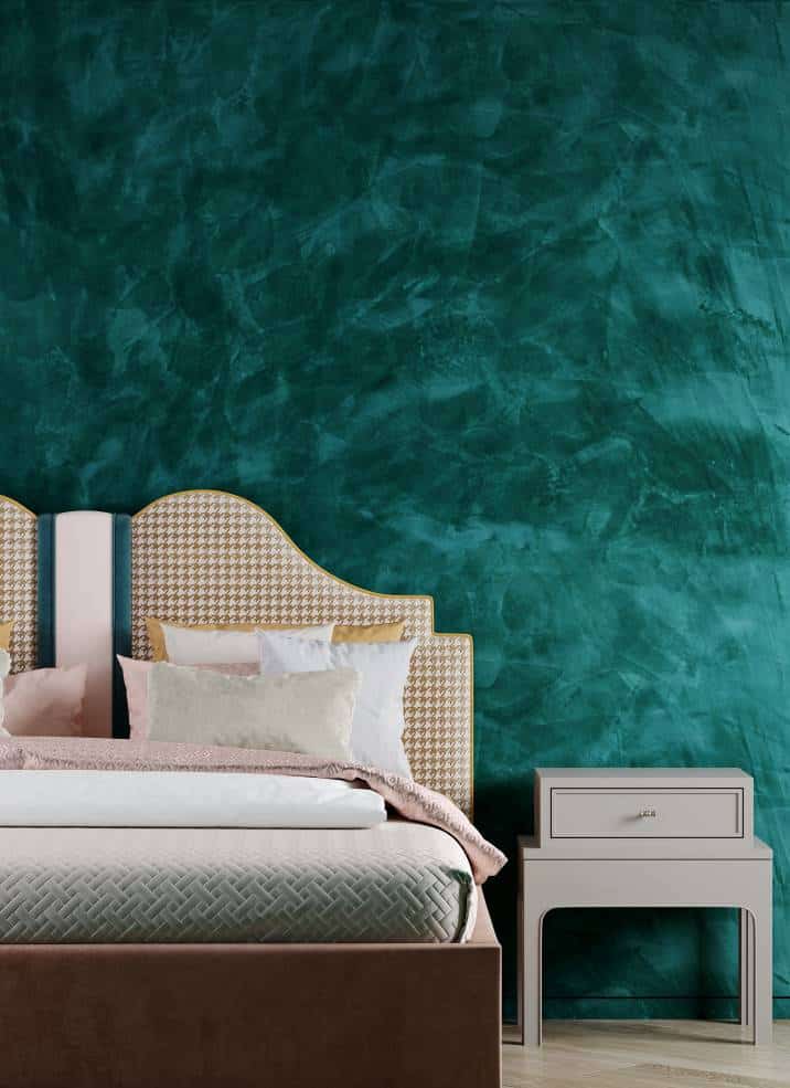 Bedroom with a green wall with stucco like smoke or waves. Textile bed in orange, pink, beige, emerald colours and houndstooth pattern. Small beige bedside table