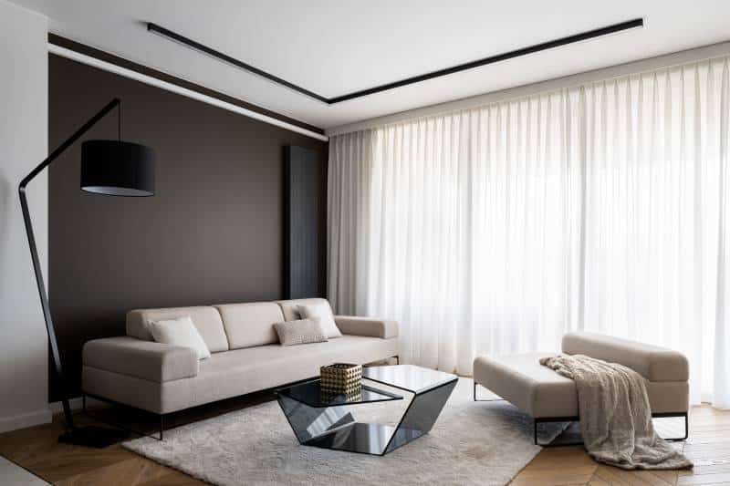 White floor-to-ceiling curtains. Simple and elegant bright living room with big windows behind white curtains. Big beige couch and seat, modern coffee table, and black lamp