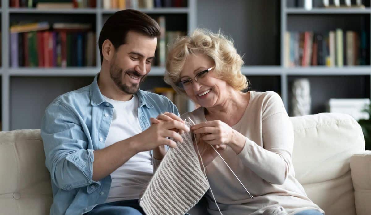 Smiling elderly mother teaching her adult son to knit using needles and yarn. Spending time together in living room on Mother’s Day