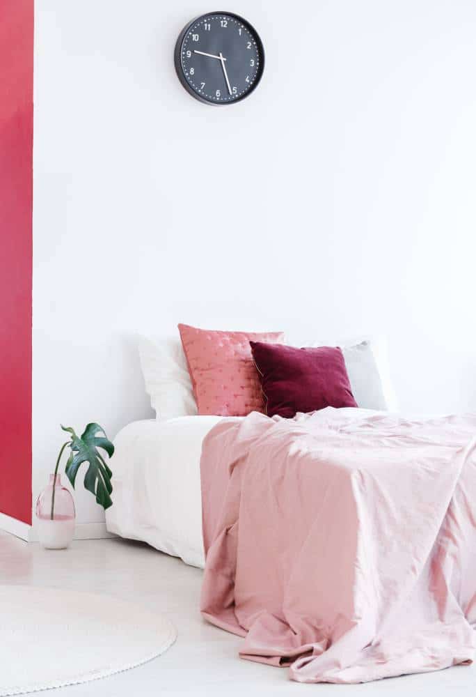Pastel pink bedsheets on bed with cushions against white wall with black clock in bedroom