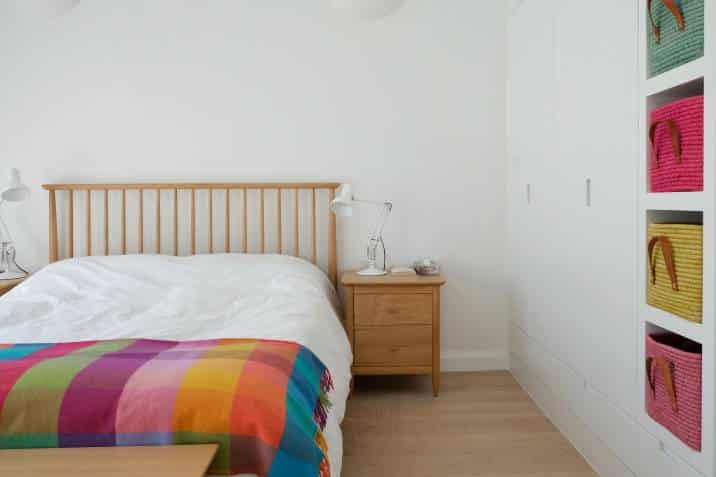 Scandinavian-inspired bedroom. Wooden bedroom furniture, white painted walls, white bedding. Colourful blanket and baskets in built-in wall shelves