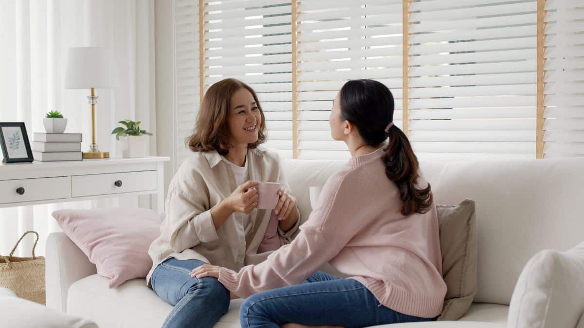 Middle-aged mother smiling and talking to grown up daughter, spending quality time in living room