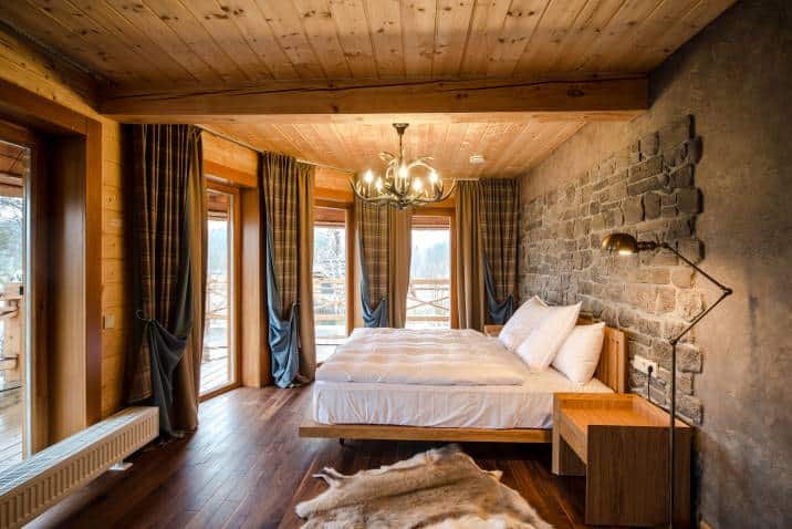 Bedroom with stonework wall. Wooden walls animal skin on floor. Lighting equipment hang on ceiling. Bed with clean, comfortable bedding blanket soft pillows. Country landscape through window