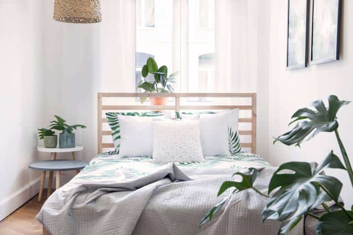 Modern Scandinavian sunny bedroom with plants, floral pattern bedding and pillows. Space with white walls and brown wooden parquet