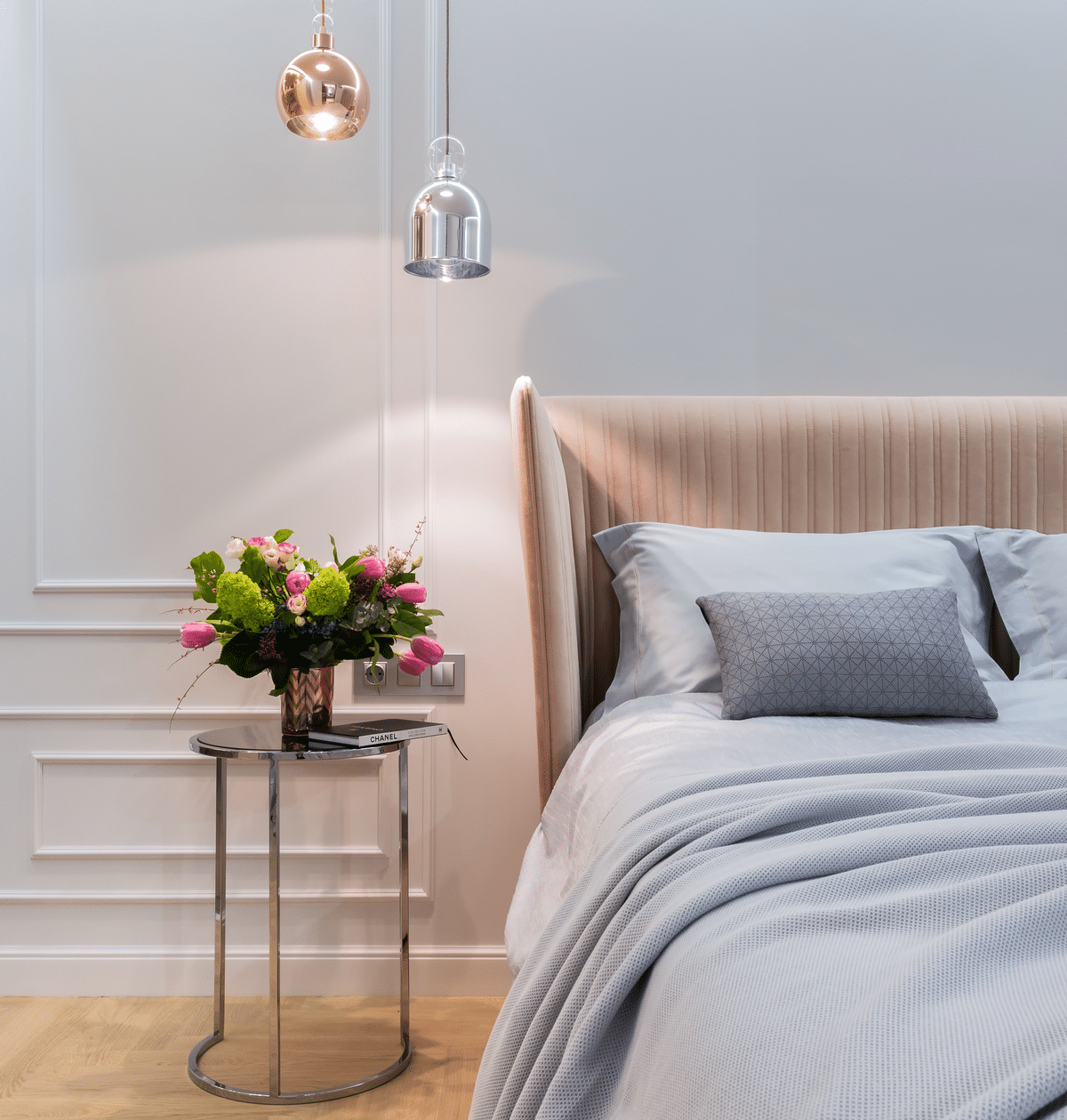 bed with grey linen and pillows next to a small table with gold and silver lights suspended above it