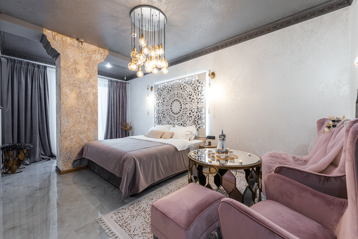 Bedroom with a wide marble column and bed with intricate headboard. A high ceiling with a hanging sphere chandelier