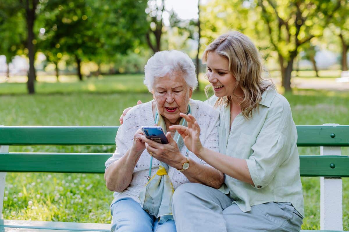 Adult granddaughter helping her grandmother to use smartphone while sitting on bench in park on Mother’s Day