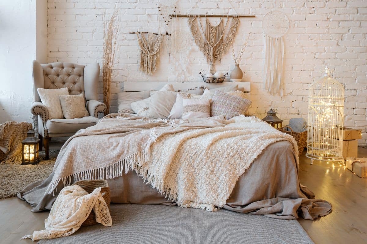 Front view of cosy Bohemian-style bedroom with fairy lights inside birdcage decor and indoor street lamp. Soft plaid and warm blanket on comfortable bed, pillows, cushions, armchair