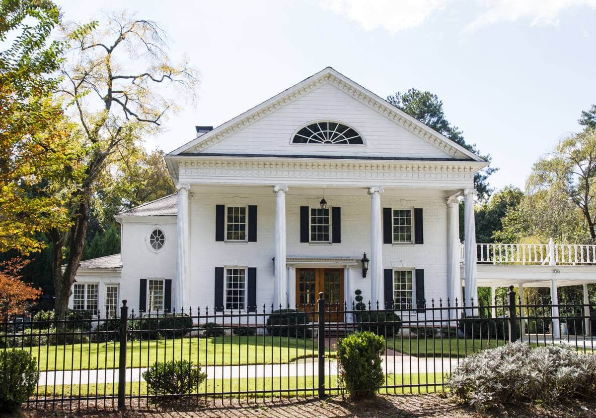 a traditional white home with columns and carport in a landscaped lawn behind a black wrought iron fence, balcony on top of carport