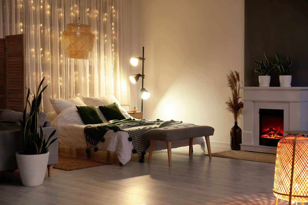 Cosy bedroom with curtain fairy lights behind bed. Modern floor lamp, houseplants, and fireplace in evening