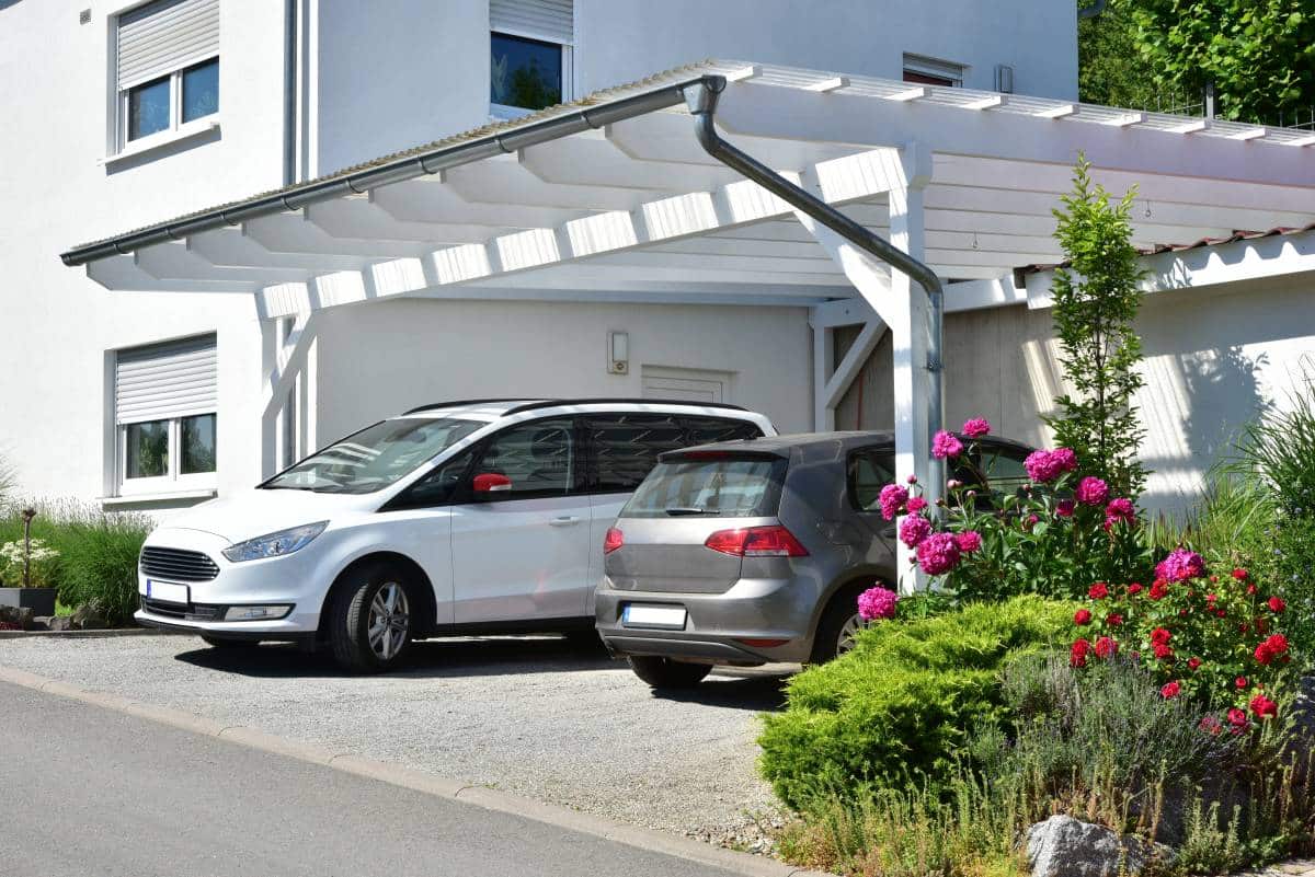 White wooden carport with two cars parked under it. The carport is beside a residential building. 