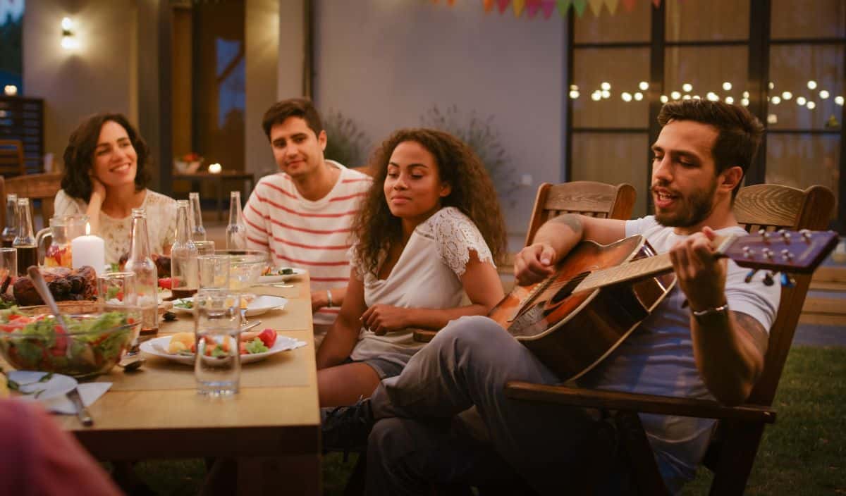 Family sitting at dinner table outdoors, young man plays the guitar to sing a personalized song for his mom on Mother’s Day