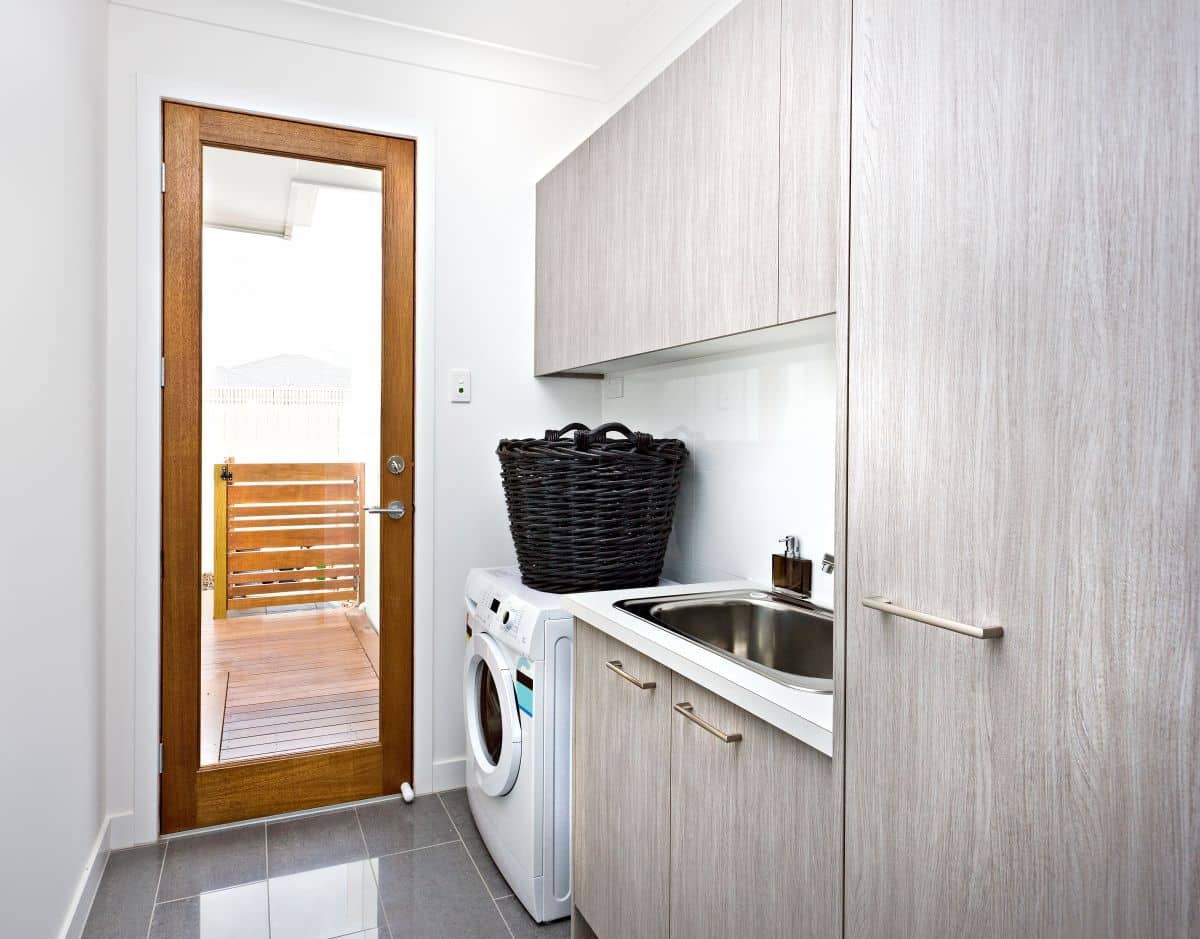 Minimalist laundry room with grey cupboards on the wall near wooden door with glass panel closed. A black rattan basket sits on the washing machine.