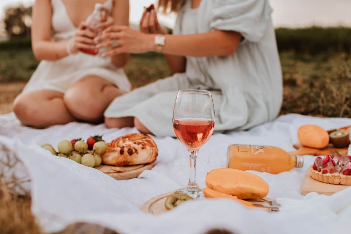 Fruits, cheese, wine, and pastries on picnic blanket. Mother and daughter sitting in the background
