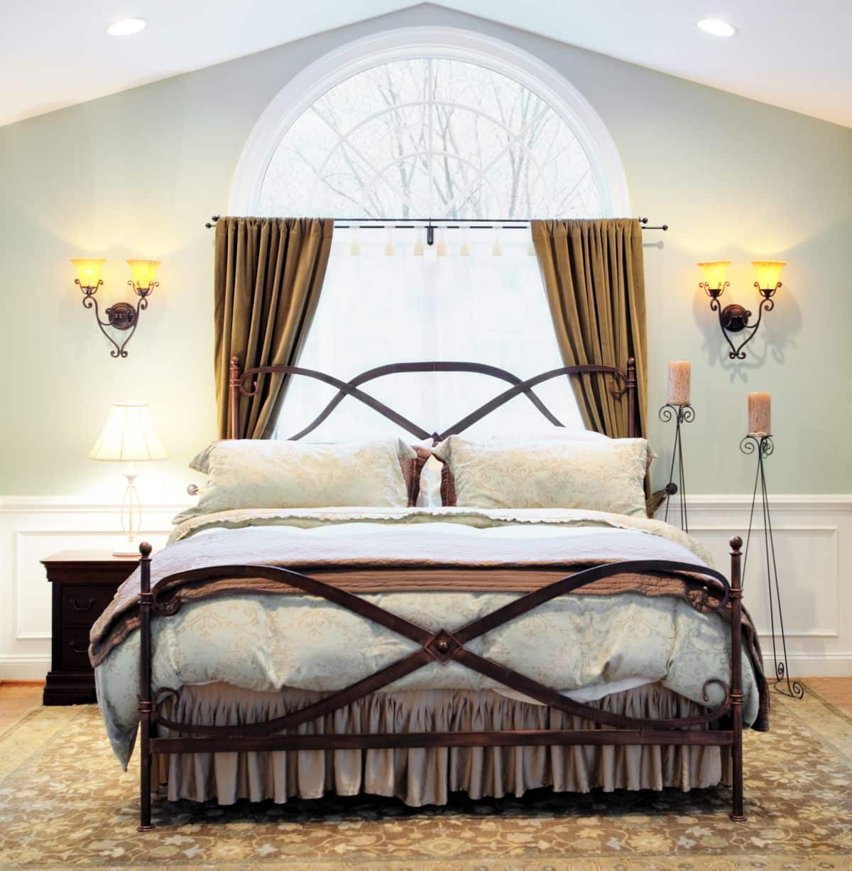 Upscale bedroom with wall torch lights on both sides of the bed. Arched window, vaulted ceiling, and ornate metal bed frame