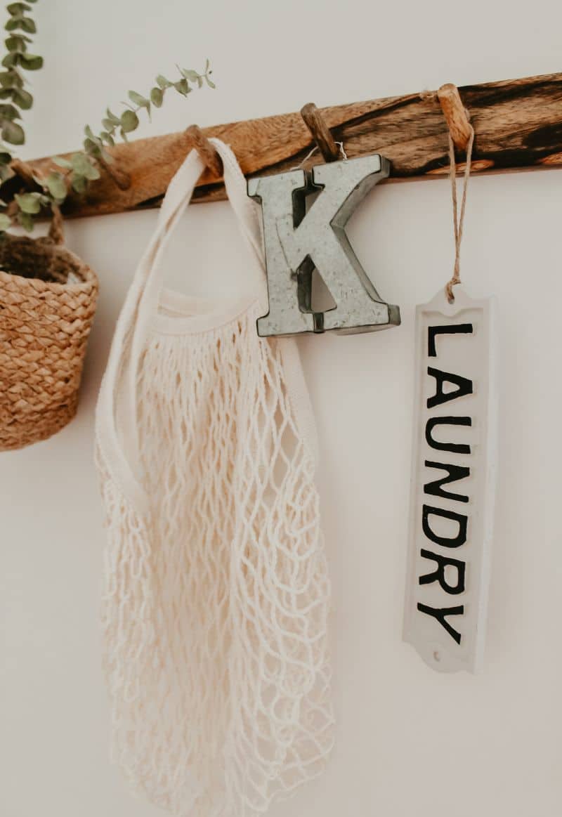 Closeup of farmhouse wall hanger in laundry room. Crochet tote bag, letter “K” keychain, “laundry” tag keychain, and eucalyptus in a basket hanging from the hooks 