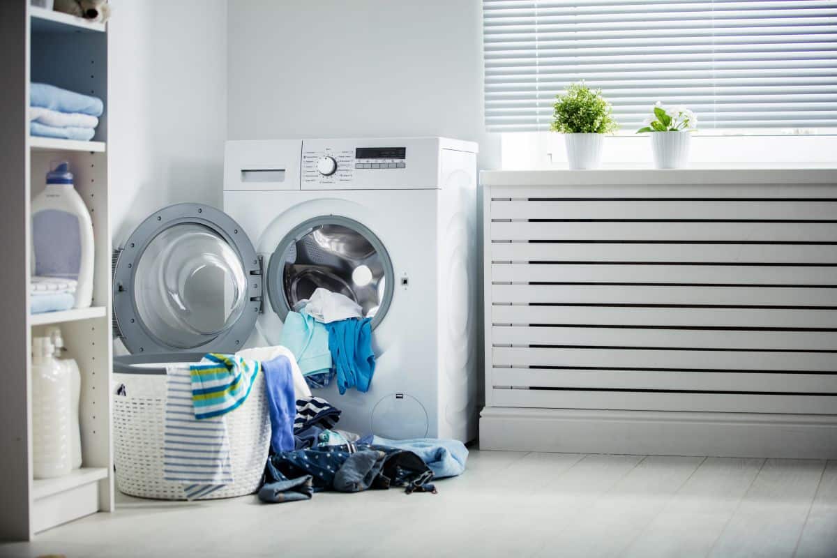 White and sleek dresser beside washing machine. Pile of dirty clothes in washing machine and adjacent basket 