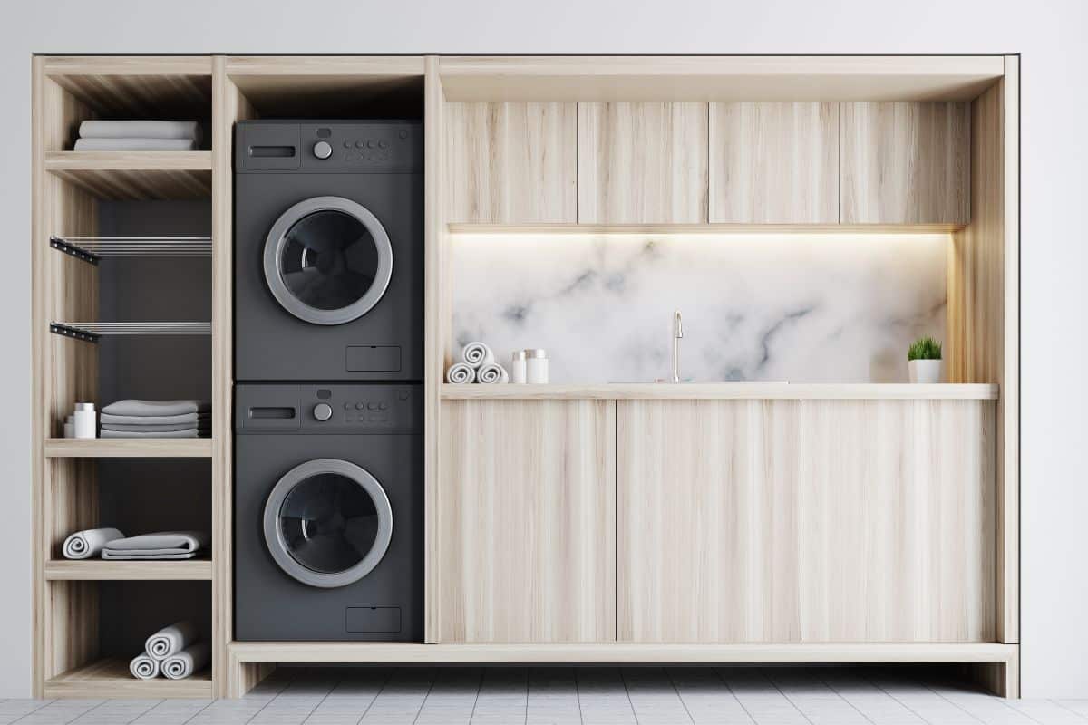 Sleek, wooden cabinets and shelf in laundry room. Marble wall, built-in washing machines, and under-cabinet lighting 