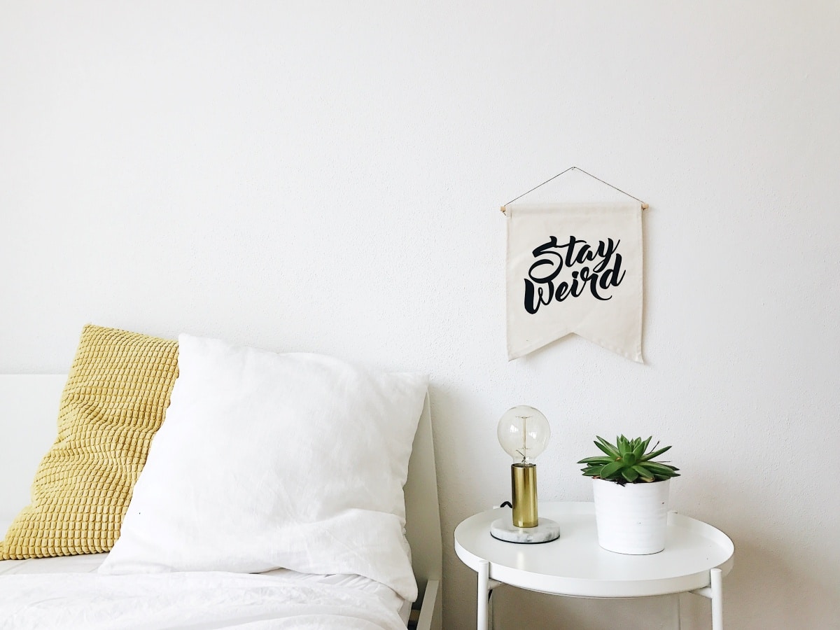 Bed with a potted plant on bedside table and a "stay weird" banner on the wall