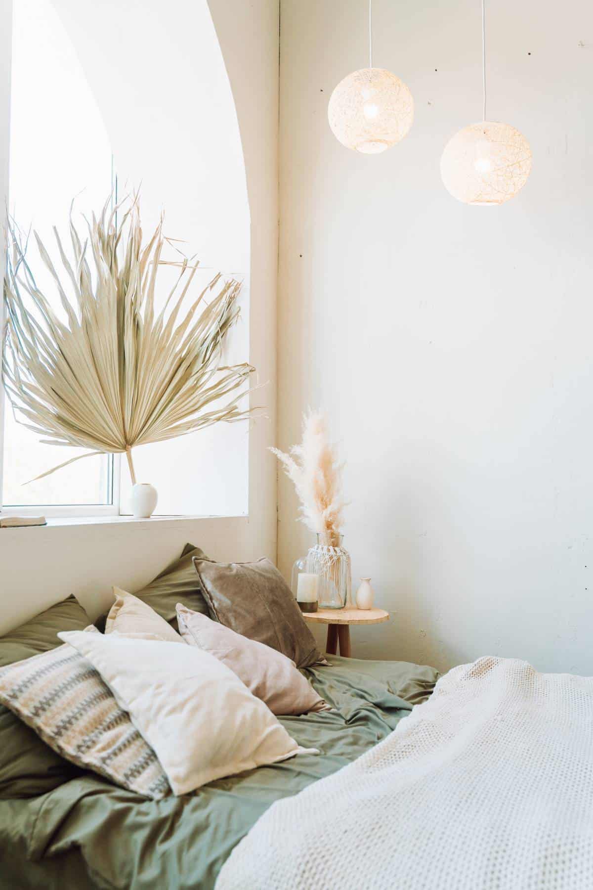 Interior of sunny modern bedroom with bed covered with grey bed linen, beige plaid and a huge palm leaf in white vase on windowsill.