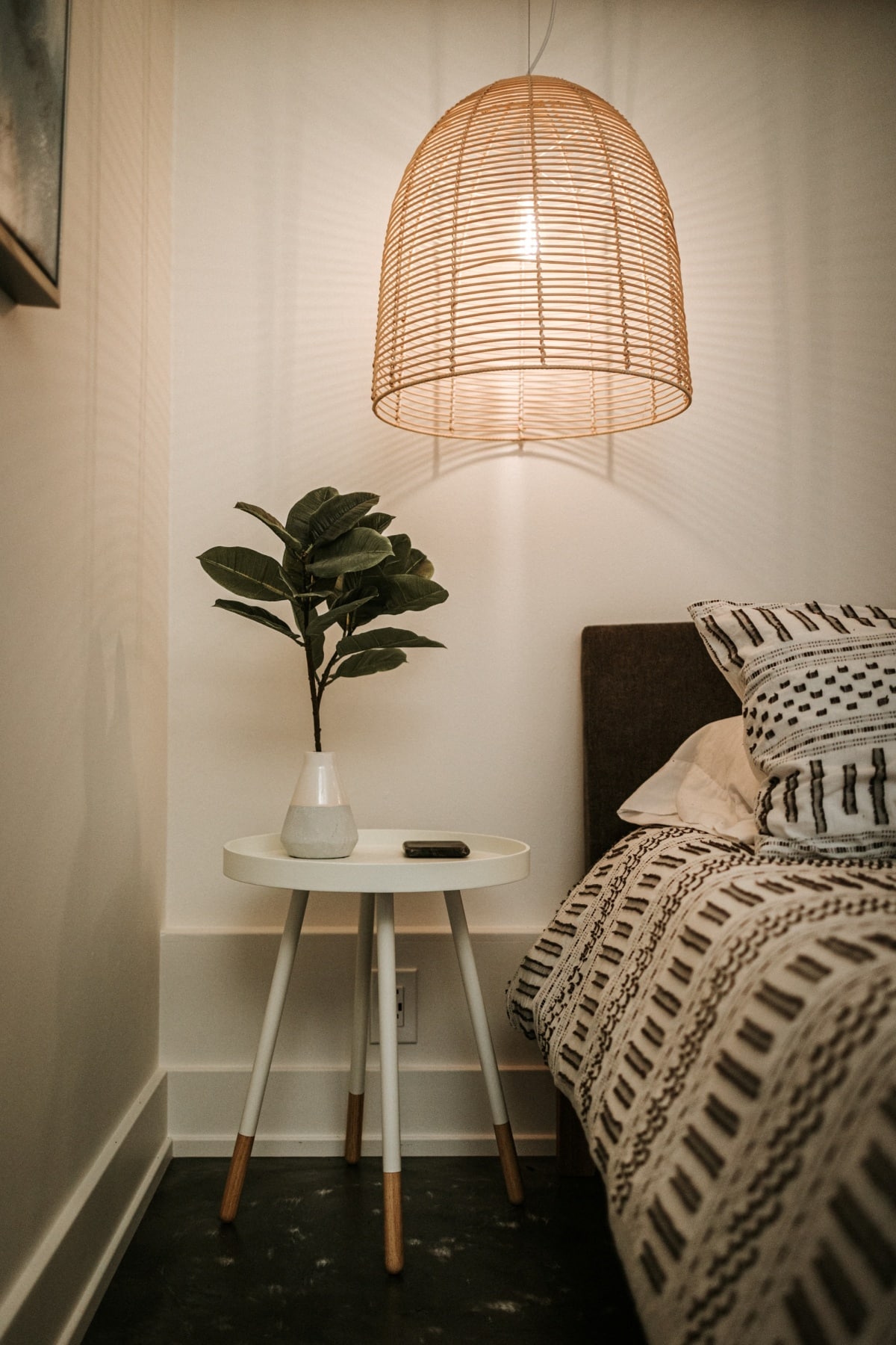 A basket hanging lamp above a white bedside table with plant