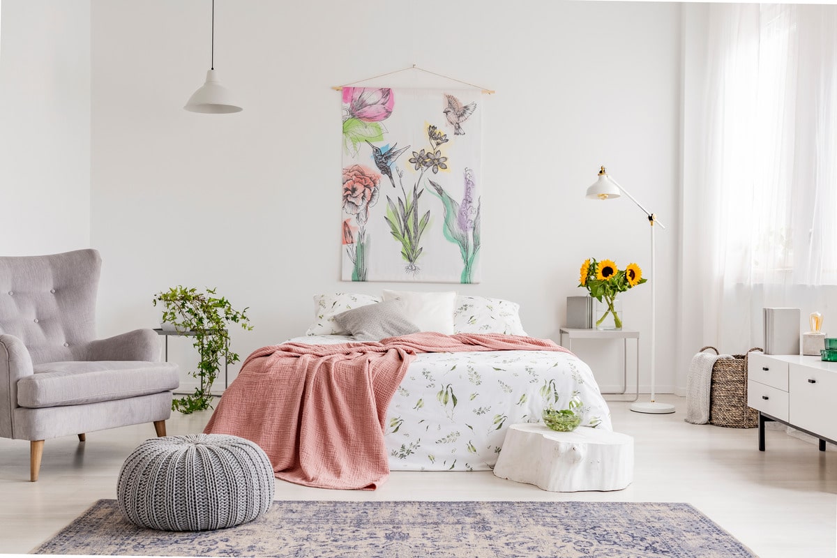 bright bedroom interior with a wall art of flowers and birds painted on a fabric above a bed which is dressed in green plants pattern on white linen