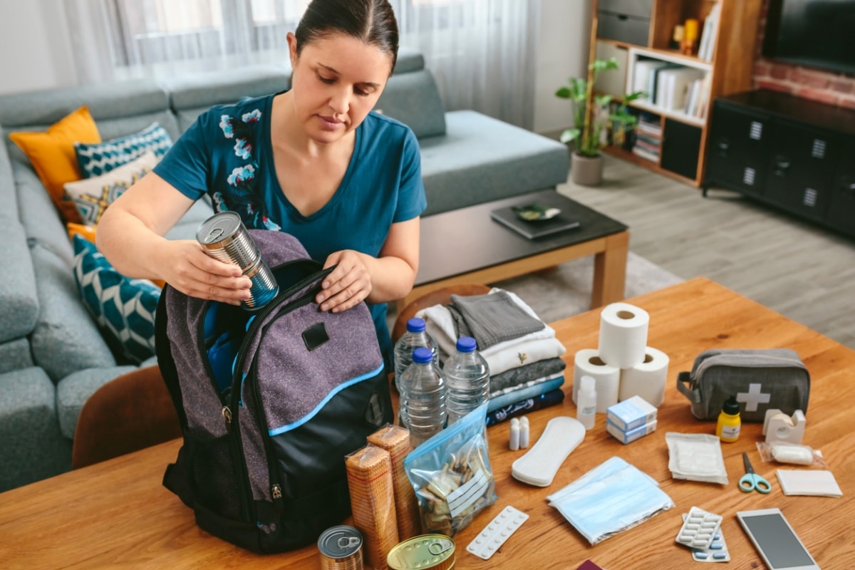 A woman preparing an emergency kit, which includes canned food, bottles of water, extra clothes, medication, and personal hygiene items.