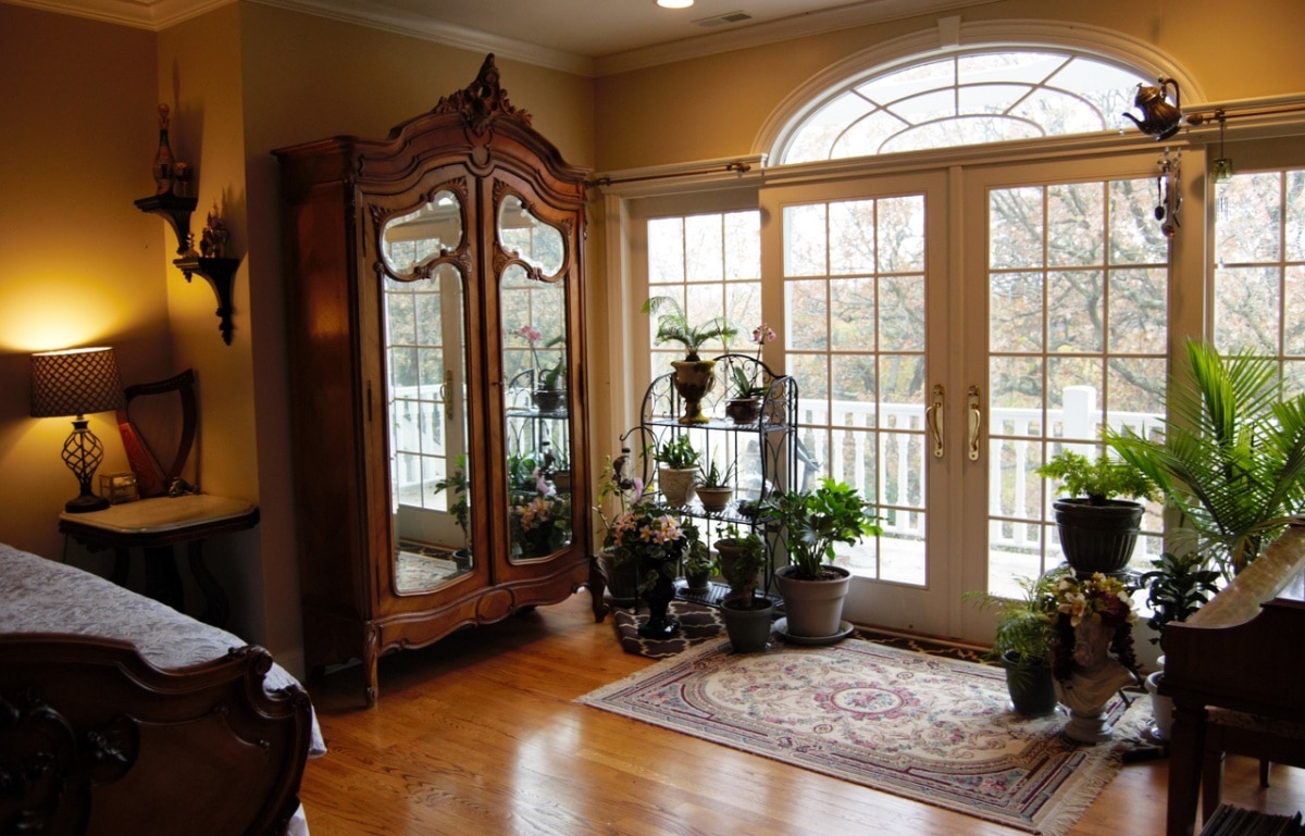 A bedroom with French doors out to a balcony, antique armoire, and houseplants