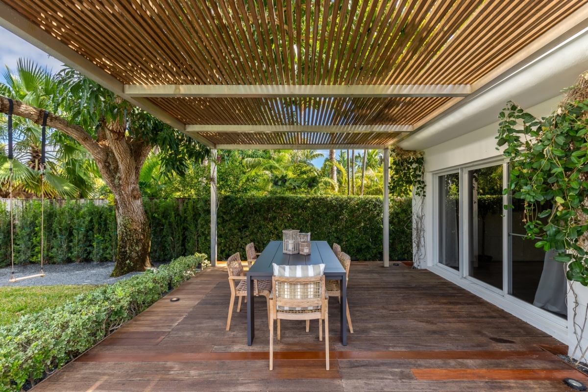 A backyard with a wooden deck and a dining table. The table has chairs and a vase with flowers. There are plants and a house in the background.