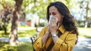 A woman in a mustard-yellow jacket sneezing into a tissue while outdoors in a park